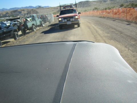 1990 Ford Bronco - View over a vehicle hood down a dirt pit road lined with parked trucks and orange fencing in the desert.