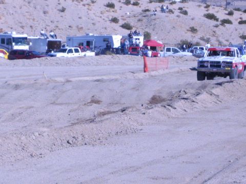 1990 Ford Bronco - off-road race truck rounds a dirt track as spectators watch from RVs and pit vehicles in the desert.
