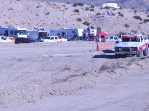 1990 Ford Bronco - white and red off-road race truck corners on a sandy dirt track lined with RVs and spectators.
