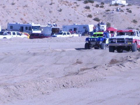 1990 Ford Bronco - green-and-blue race truck races alongside a red and white truck on a sandy desert course.