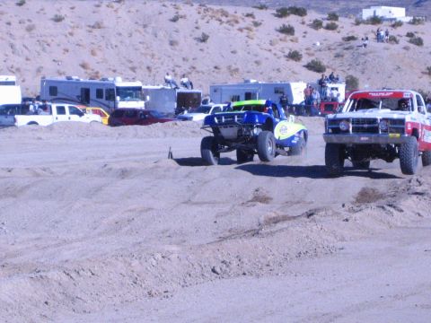 1990 Ford Bronco - blue race truck and a red truck crest a sandy berm side by side on a desert dirt course.