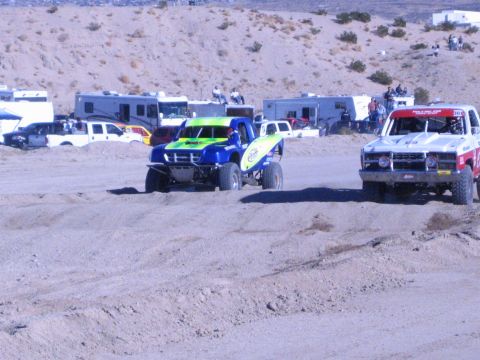 1990 Ford Bronco - blue-green race truck and a red and white truck race across a sandy desert track near the pits.