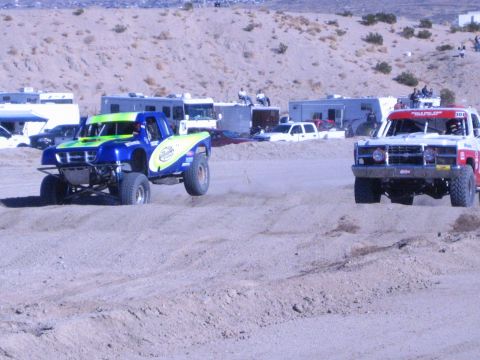 1990 Ford Bronco - green-and-blue off-road race truck runs near a red and white truck on a sandy desert course.