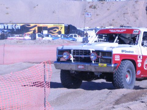 1990 Ford Bronco - Front view of a white-and-red number 301 off-road truck with round headlights crossing a dirt track lined with orange fencing.