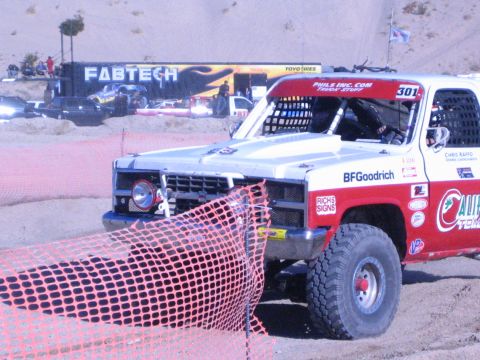 1990 Ford Bronco - number 301 off-road race truck rounds a dirt course as orange safety fencing droops in front and a FABTECH trailer sits behind.