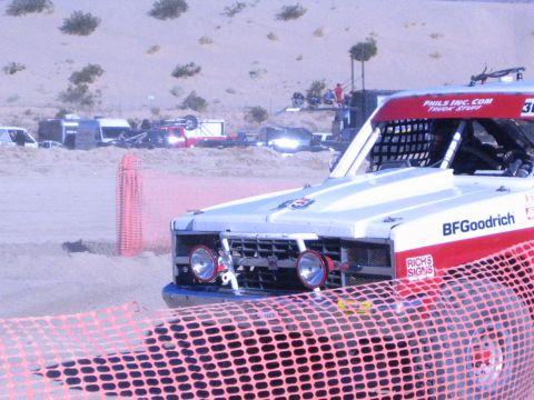 1990 Ford Bronco - Close front view of a white-and-red off-road race truck with twin round headlights behind sagging orange safety fencing in the desert.