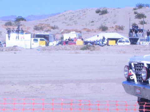 1990 Ford Bronco - wide desert pit scene with trailers, tents and crowds under blue sky, the nose of a race truck visible at lower right behind orange fencing.