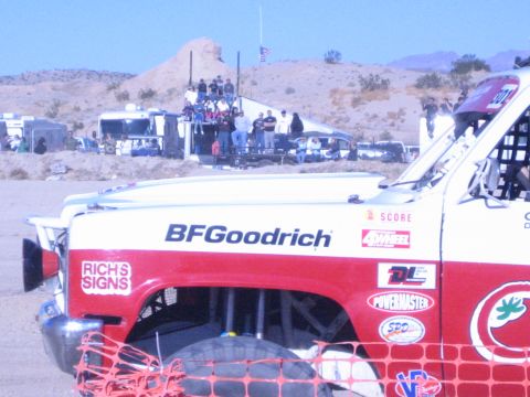 1990 Ford Bronco - Side view of a red-and-white off-road race truck with BFGoodrich and sponsor decals, spectators watching from a platform in the desert beyo…