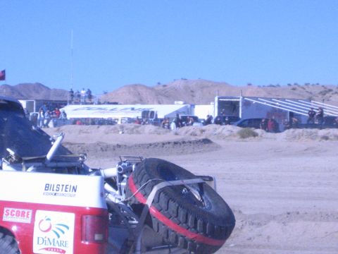 1990 Ford Bronco - Rear quarter of a red-and-white off-road race truck with a bed-mounted spare tire, facing a desert pit row of trailers and spectators.