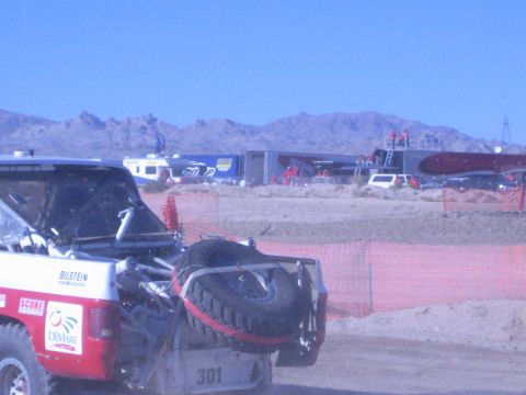 1990 Ford Bronco - Rear view of the number 301 race truck with a mounted spare tire behind orange fencing, desert pits and distant mountains in the background.