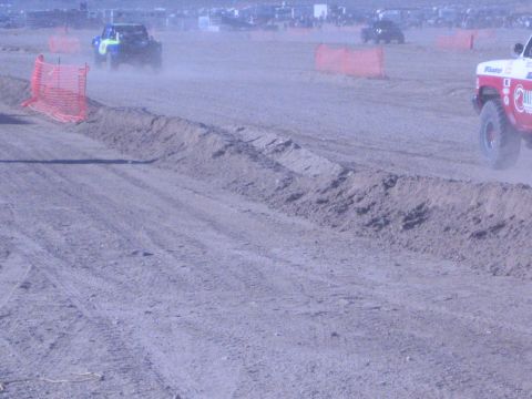 1990 Ford Bronco - Two off-road race vehicles kick up dust on a wide bermed dirt course marked by orange fencing, one red-and-white truck at right.