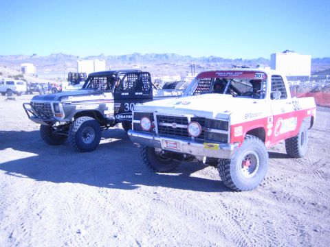 1990 Ford Bronco - silver vintage-bodied off-road truck and the red-and-white number 301 truck parked side by side in a desert staging area.
