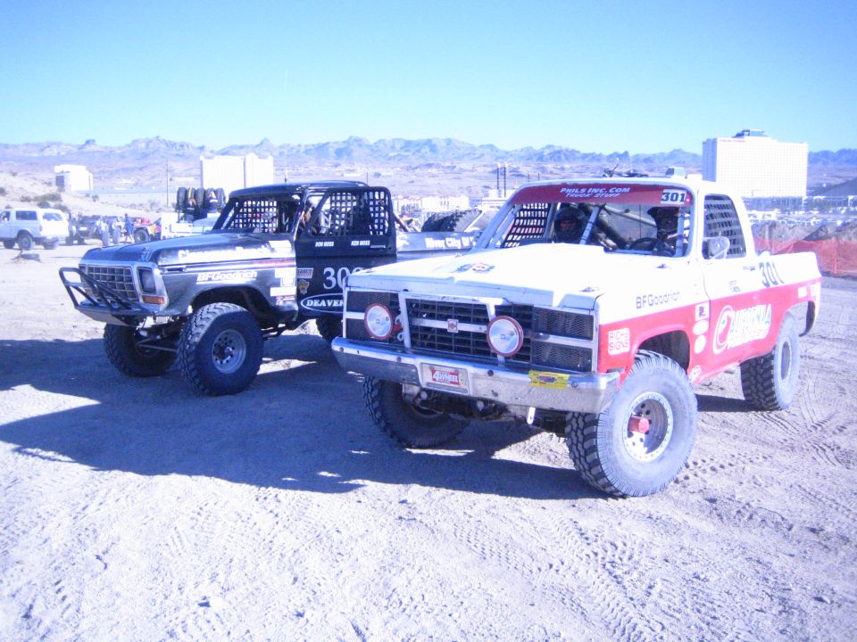 Two off-road trucks parked side by side in a desert pit, one silver, one red and white number 301