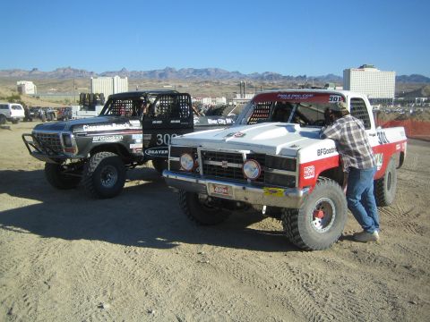 1990 Ford Bronco - silver truck and the red-and-white number 301 truck parked in a desert staging area, a person standing beside the 301 with buildings behind.