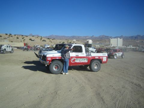 1990 Ford Bronco - person leans against the side of the white-and-red number 301 off-road truck parked in a dirt lot with mountains and buildings beyond.