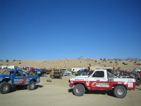 1990 Ford Bronco - field of off-road race trucks staged on the desert sand, with a blue Horizon Realty truck and the red-and-white number 301 in front.