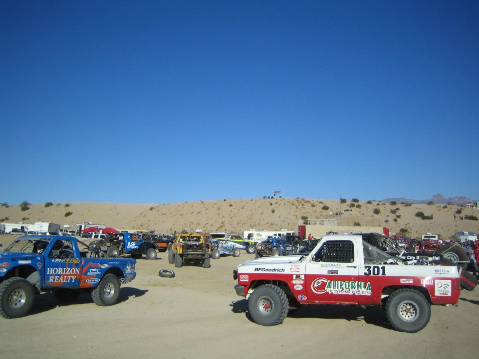 Off-road race trucks staged in the desert, a blue Horizon Realty truck and red-white number 301 in foreground