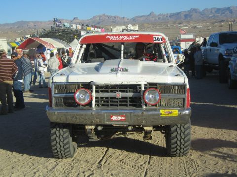1990 Ford Bronco - Front three-quarter view of the white-and-red number 301 off-road truck in a crowded desert pit area with tents and spectators.