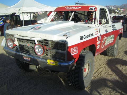 1990 Ford Bronco - Front of the white-and-red number 301 off-road race truck with twin round headlights, surrounded by canopy tents in a desert pit.