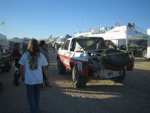 1990 Ford Bronco - Rear view of the red-and-white number 301 off-road truck with a bed-mounted spare in a bustling desert pit as spectators walk past.