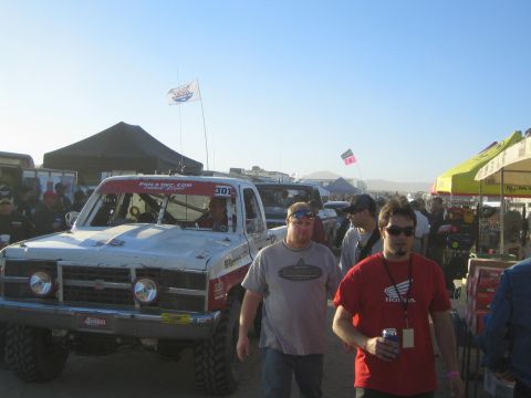 1990 Ford Bronco - number 301 off-road truck sits in a busy pit at low sun while two men walk in front, one wearing a red Honda T-shirt.