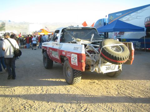 1990 Ford Bronco - Rear three-quarter view of the white-and-red number 301 off-road truck with a mounted spare in a busy desert pit under blue tents.