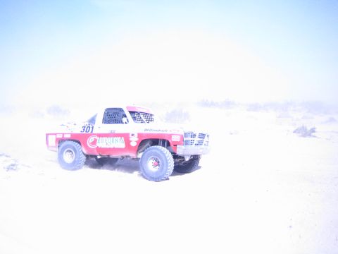 1990 Ford Bronco - red-and-white number 301 off-road race truck moves across pale, dusty desert terrain in bright washed-out light.