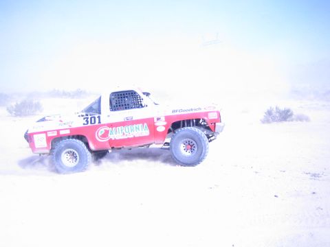 1990 Ford Bronco - red-and-white number 301 off-road truck speeds across desert sand, throwing up a trail of dust under a hazy sky.