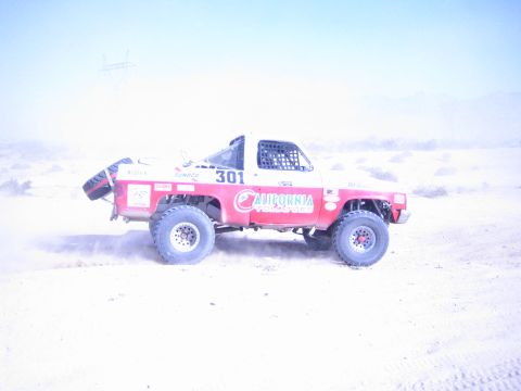 1990 Ford Bronco - red and white off-road race truck marked 301 with California sponsor graphics speeds through a dusty desert course.