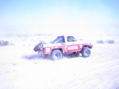 1990 Ford Bronco - Off-road race truck number 301 races across sandy desert terrain, trailing a plume of dust behind it.