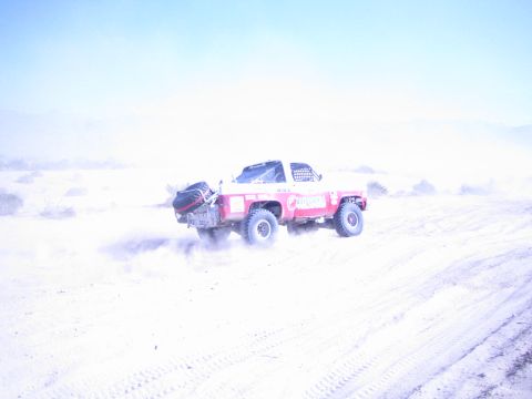 1990 Ford Bronco - red and white off-road race truck powers over a sandy desert rise, spraying dust under blue sky.