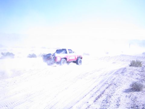 1990 Ford Bronco - red and white off-road race truck climbs a sandy desert berm amid a wash of dust and bright haze.