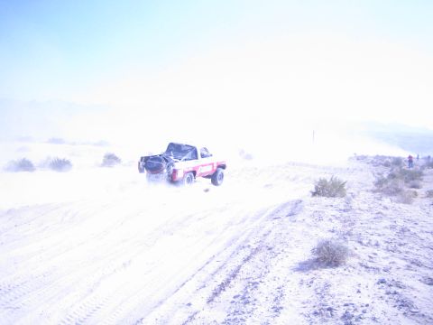 1990 Ford Bronco - red and white race truck drives a sandy desert trail as faint spectators stand on a distant ridge.