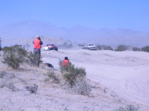 1990 Ford Bronco - Spectators in orange safety vests watch two off-road race trucks navigate dusty desert hills under mountains.