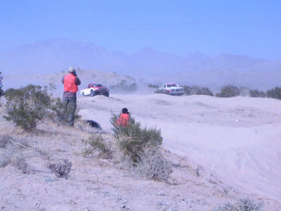Spectators in orange vests watch two race trucks across the desert