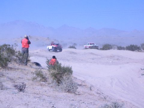 1990 Ford Bronco - People in orange vests photograph two off-road race trucks crossing sandy desert terrain backed by mountains.