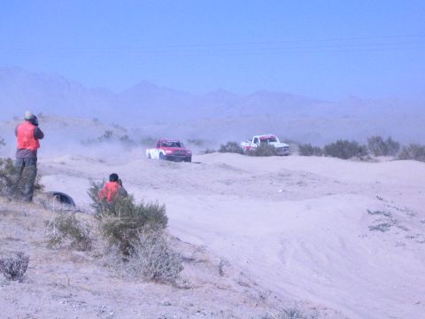 1990 Ford Bronco - red truck and a white truck race across a sandy desert course as orange-vested spectators look on.