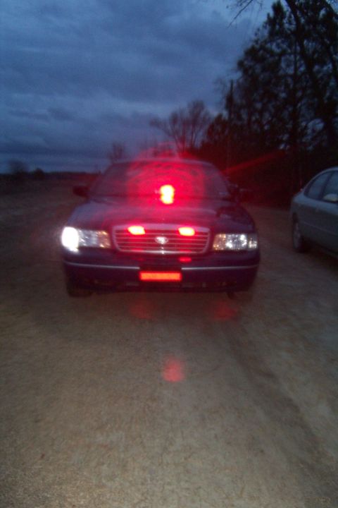 1998 Ford Crown Victoria - Head-on night shot of a dark Ford Crown Victoria sedan on a dirt lot at dusk with its emergency lighting running: a red rotating beacon on…