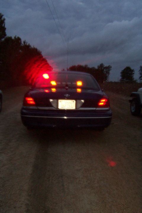 1998 Ford Crown Victoria - Rear view of a dark blue Ford Crown Victoria sedan on a dirt road at dusk with its lighting active: a red rotating beacon glowing on the re…