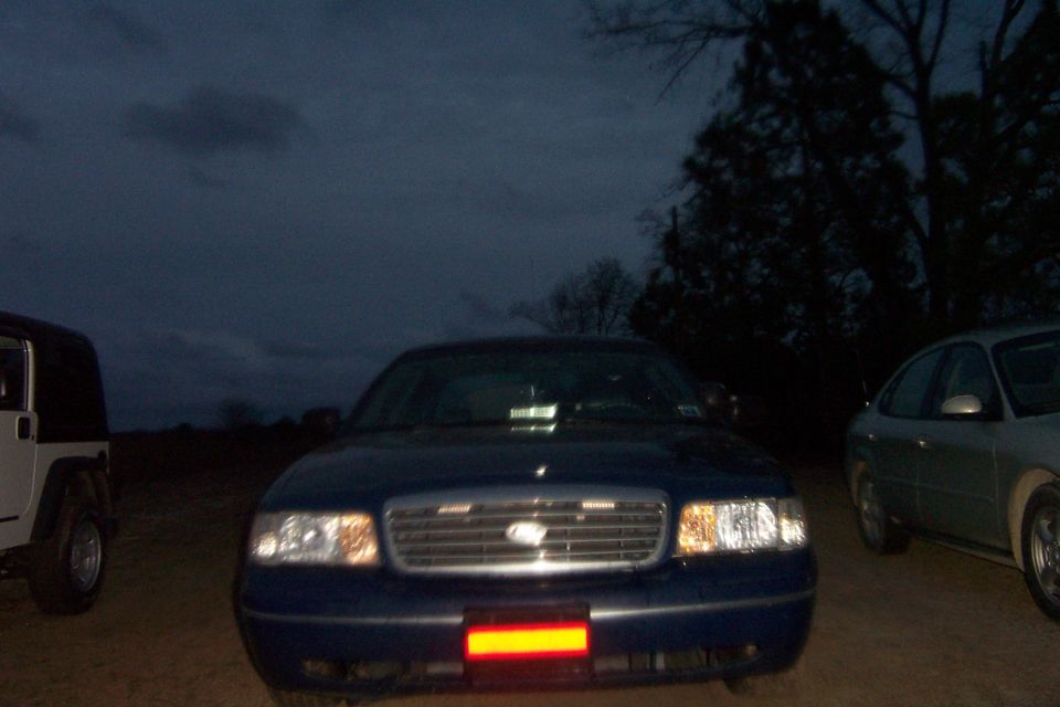Front view of a blue 1998 Ford Crown Victoria with a red light bar on the bumper, parked next to a white Jeep and a silver sedan