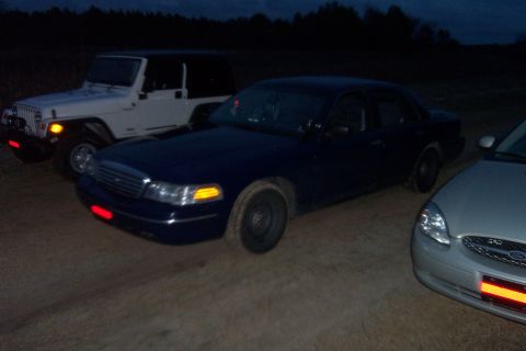 1998 Ford Crown Victoria - Three vehicles are parked on a dirt lot at dusk, with a line of trees in the background.