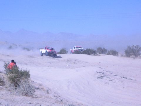 1990 Ford Bronco - red truck and a white off-road race truck crest sandy desert dunes with mountains in the hazy distance.
