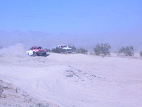 1990 Ford Bronco - Two off-road race trucks, one red and one white, traverse a sandy desert course under a blue sky.