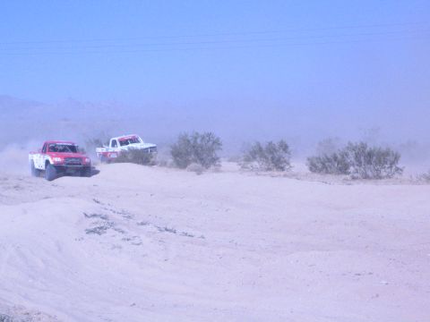 1990 Ford Bronco - red and a white off-road race truck race across pale sandy desert with mountains fading into haze.