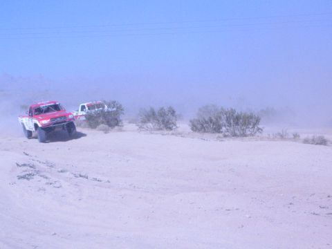 1990 Ford Bronco - red off-road race truck leads a white truck over a sandy desert rise dotted with low brush.