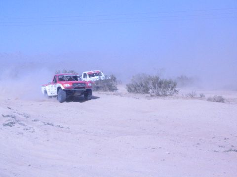 1990 Ford Bronco - red and white truck and a white truck race side by side across sandy desert terrain in a dusty haze.