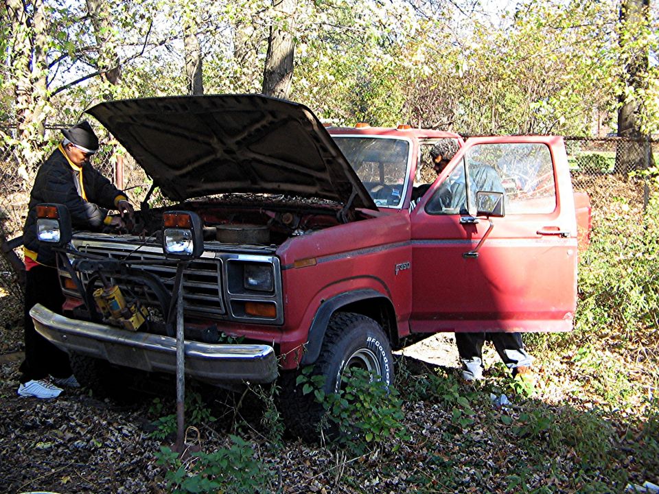 Red Ford pickup truck with hood open, someone working on engine. Door is open, another person inside