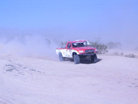 1990 Ford Bronco - red and white off-road race truck rounds a sandy desert turn, throwing up dust under a clear blue sky.
