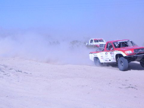1990 Ford Bronco - red and white race truck with sponsor decals leads a white truck through a dusty desert course.