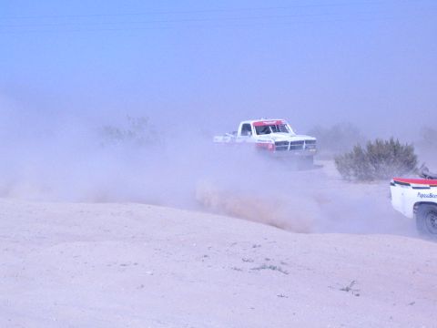 1990 Ford Bronco - white off-road race truck and a second trailing truck push through a thick cloud of desert dust.